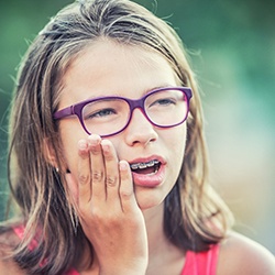 A young girl wearing purple glasses holding her cheek and experiencing soreness from her braces