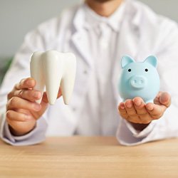 Dentist in white coat holding model tooth in one hand and blue piggy bank in the other