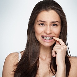 A young woman wearing traditional braces and pointing to her mouth as if she is experiencing an orthodontic emergency in McKinney